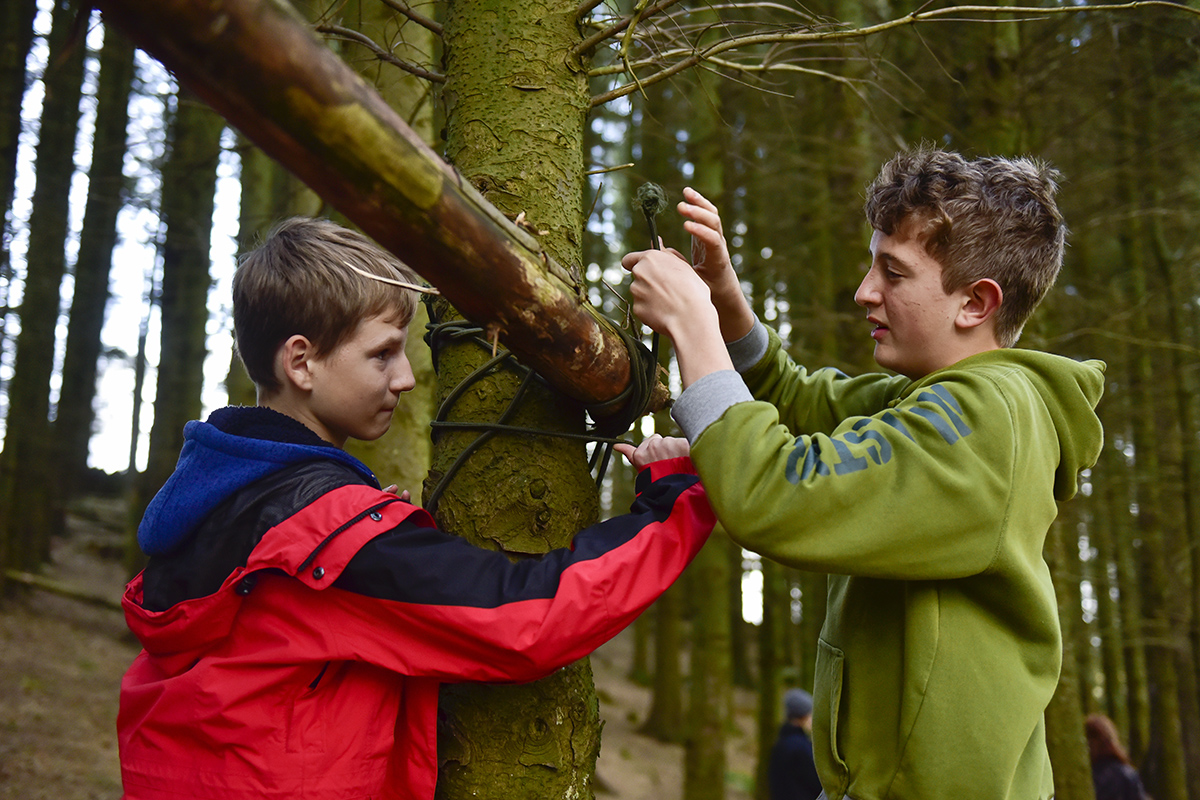 Two lads lashing a branch to a tree trunk to make a shelter