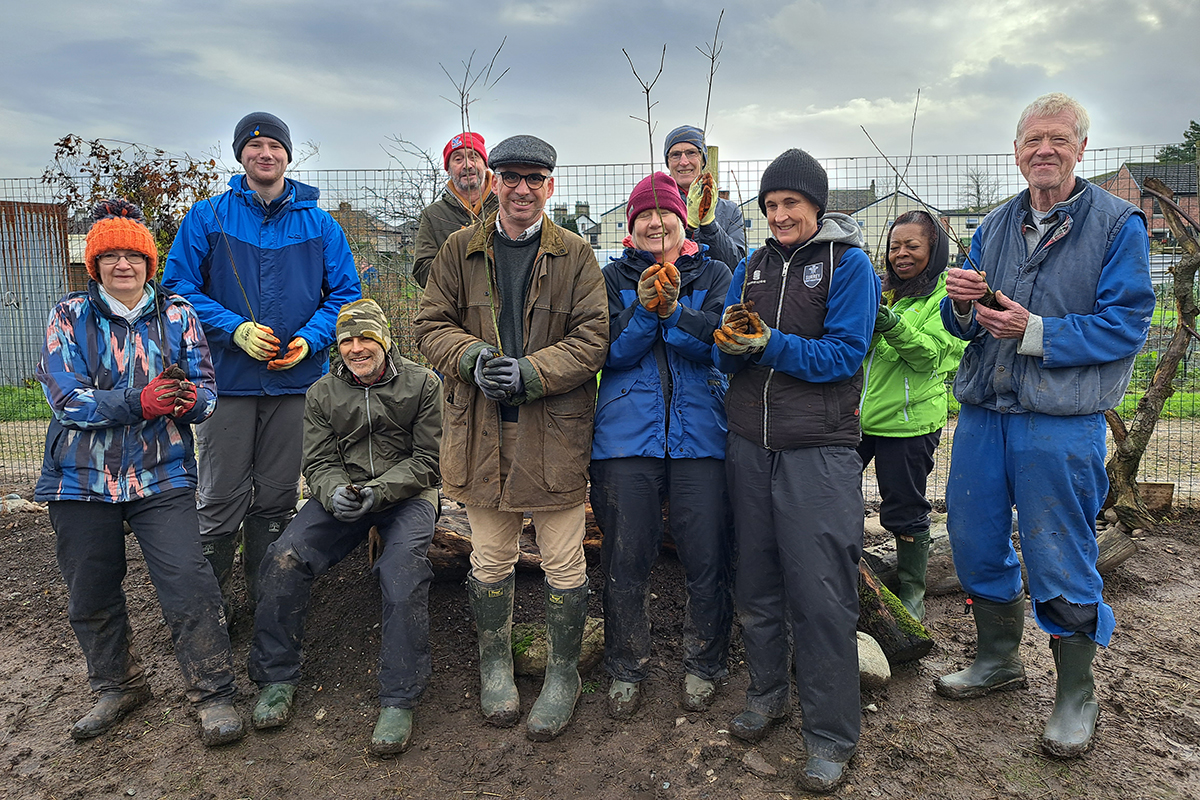 Group of people in winter coats and hats all holding plant pots containing tree whips