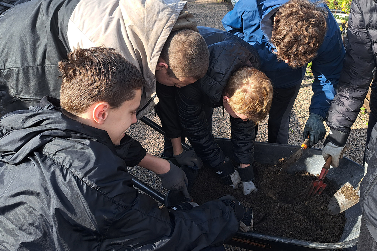 Group of boys crouched around a wheelbarrow using garden trowels,