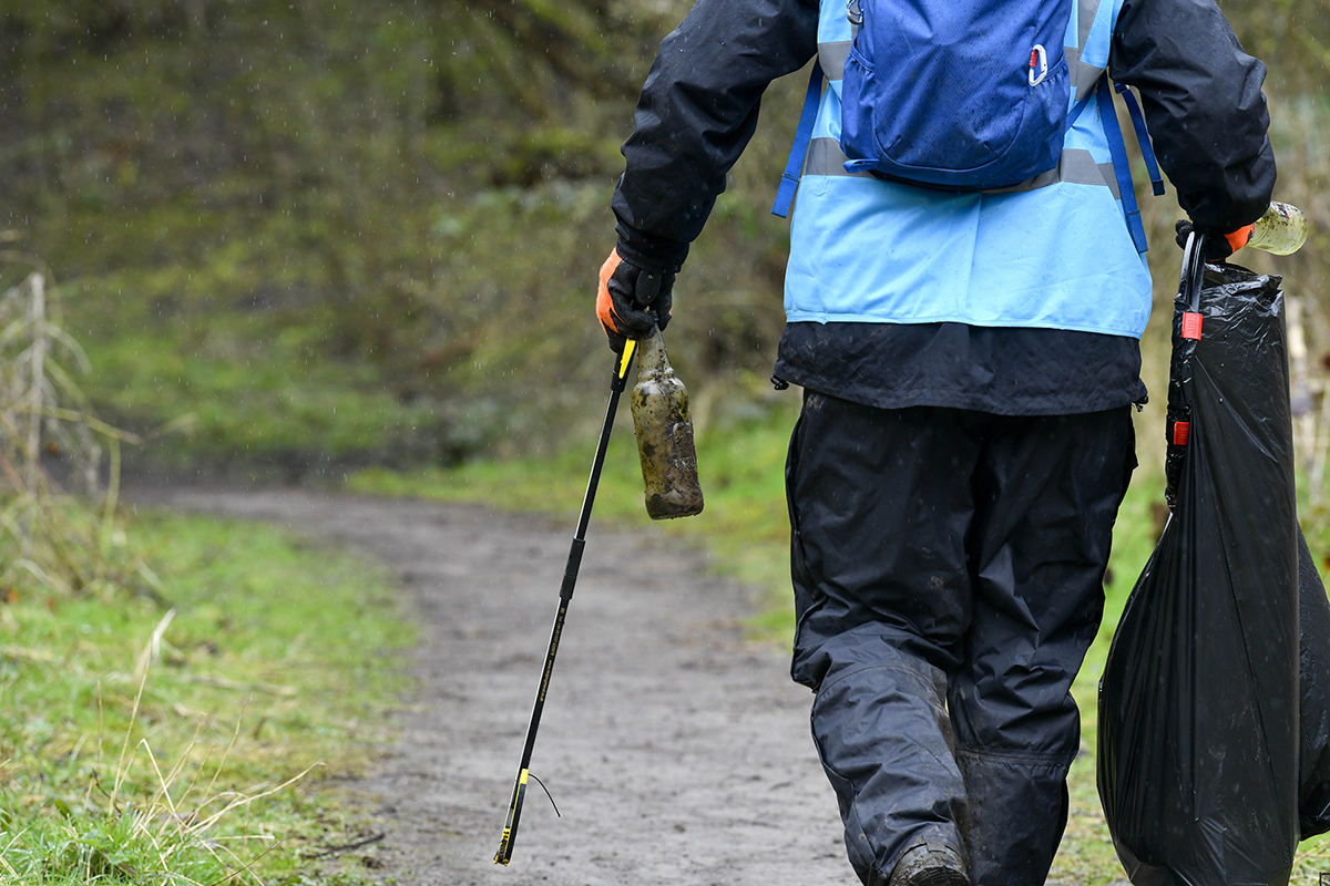 Person holding two dirty glass bottles, a bin bag and a litter picker