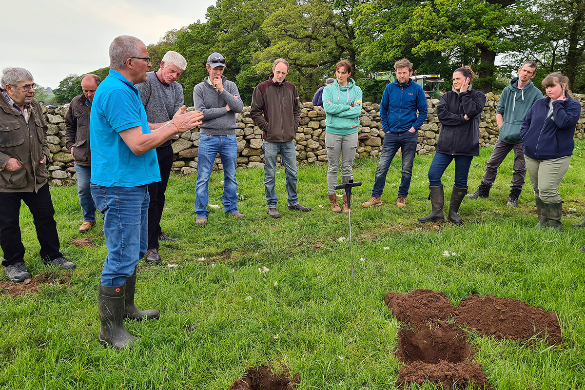 Group of people stood in a field looking at a man talking about soil in front of freshly-dug holes in the ground.