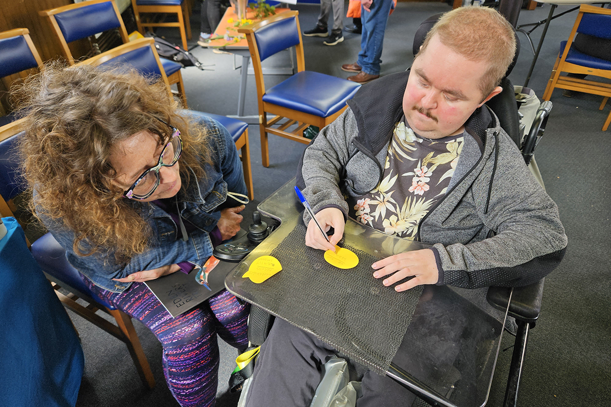 A man in a wheelchair with an attached table is writing on post it notes, whilst a woman sat next to him looks on.