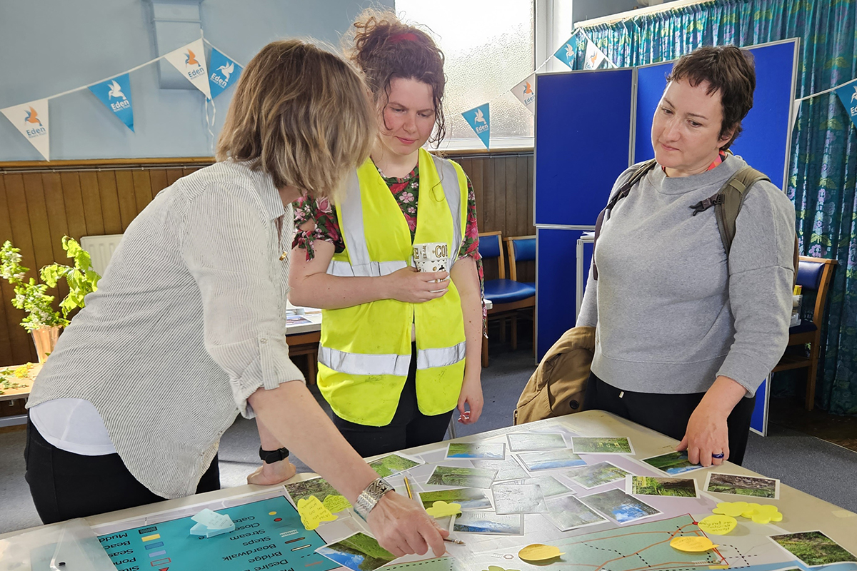 Three people stood talking next to a map on a table with post it notes stuck to it. One person is pointing at the map