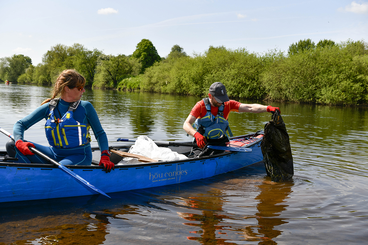 Two people sat in a canoe. The person sat at the back of the canoe is pulling a full, black bin bag out of the river.