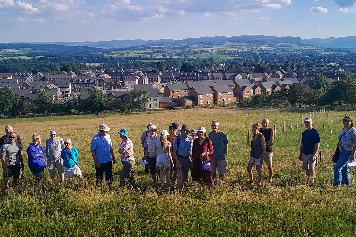A line of people in front of a field. In the background can see a housing estate and, in the distance, the Lake District fells