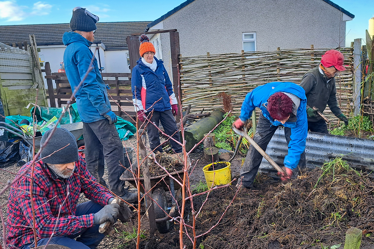 A woman uses a shovel to turn over earth on the compost heap, whilst two others look on.