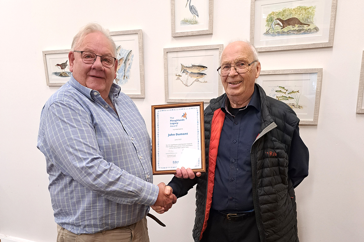 Charles Ecroyd presents a framed certificate to John Dumont. They are shaking hands and smiling at the camera.