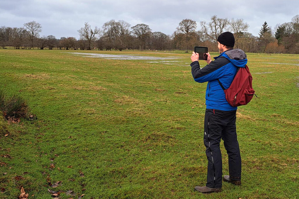 A man is stood in a field that has a pond in the corner. He is holding a tablet to take picture of the landscape.